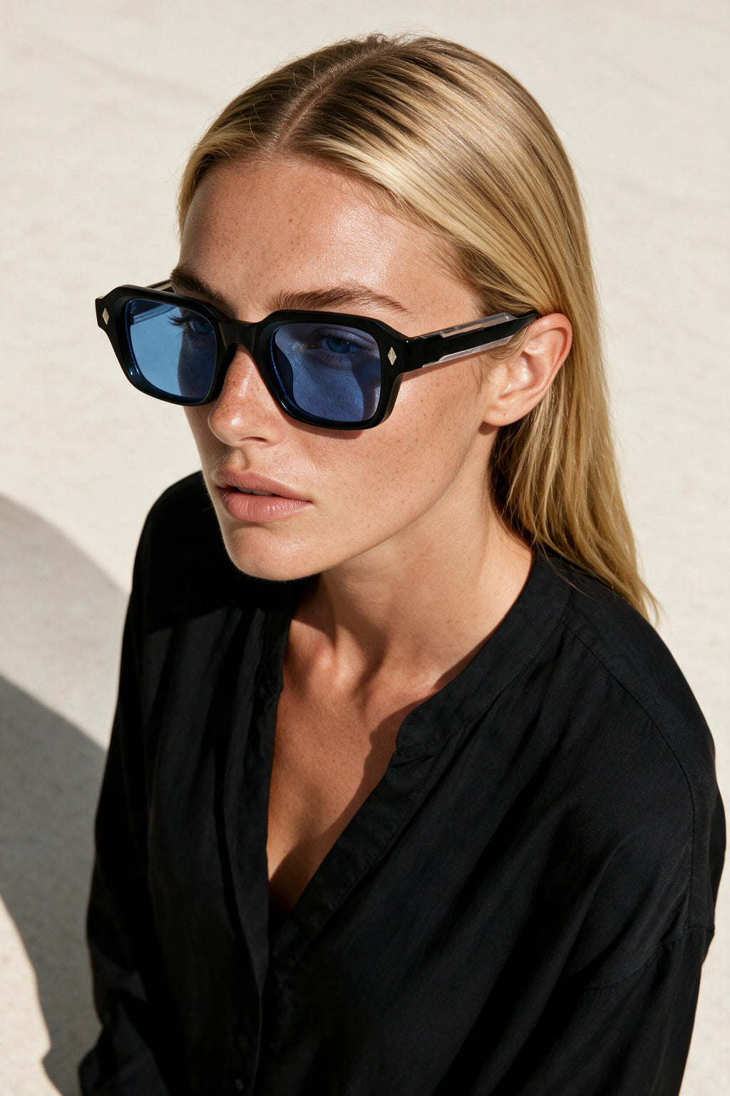 Woman wearing VELA Cobalt sunglasses, shot from a high angle against a warm bone backdrop