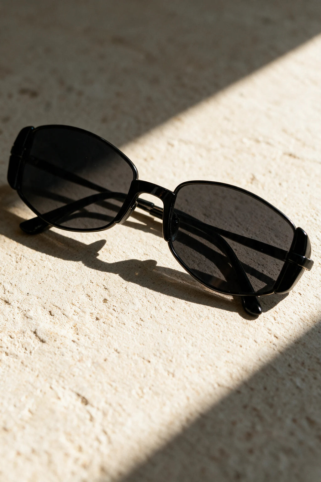 NEVA sunglasses in black folded on a warm bone plaster surface in raking sunlight