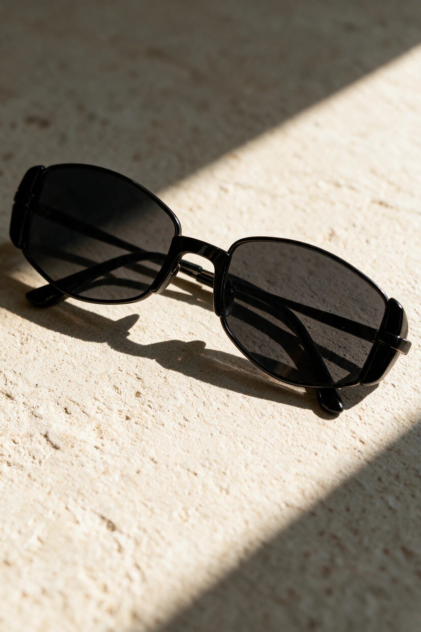 NEVA sunglasses in black folded on a warm bone plaster surface in raking sunlight