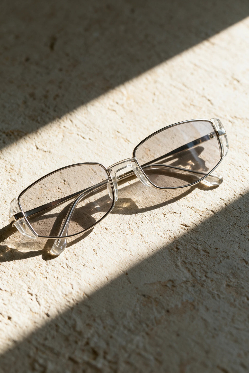 NEVA sunglasses in silver and clear crystal folded on a warm bone plaster surface in raking sunlight