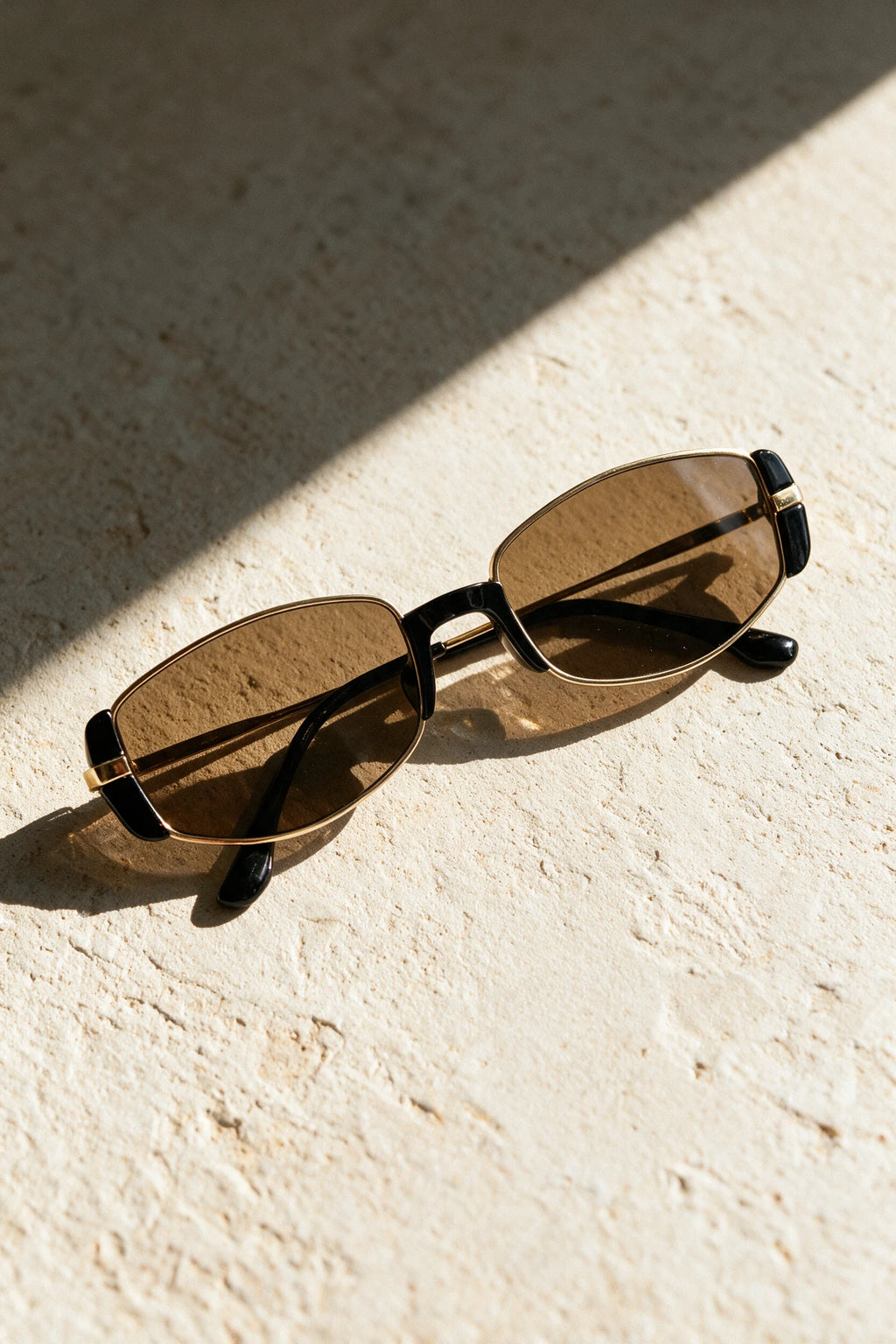 NEVA sunglasses in gold folded on a warm bone plaster surface in raking sunlight